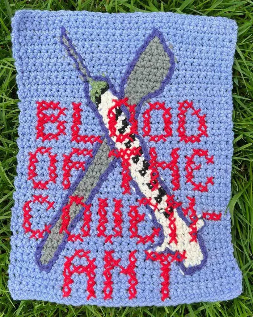 a crochet tapestry with a light purple background. There is a scalpel and syringe crossed over each other forming an X. Cross stitched over them is the phrase "BLOOD OF THE COVENANT"