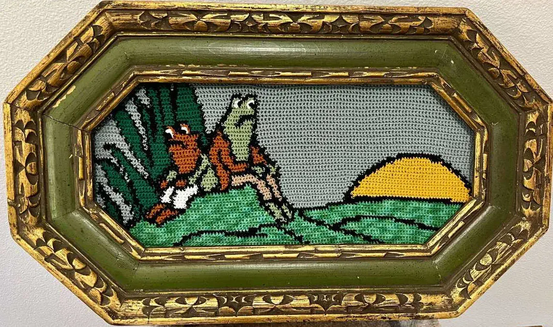 a crochet tapestry with frog and toad in the left forefront back to back, some foliage behind them. To the right is a sunset over the hills. Tapestry is in an ornate green frame with gold detailing.