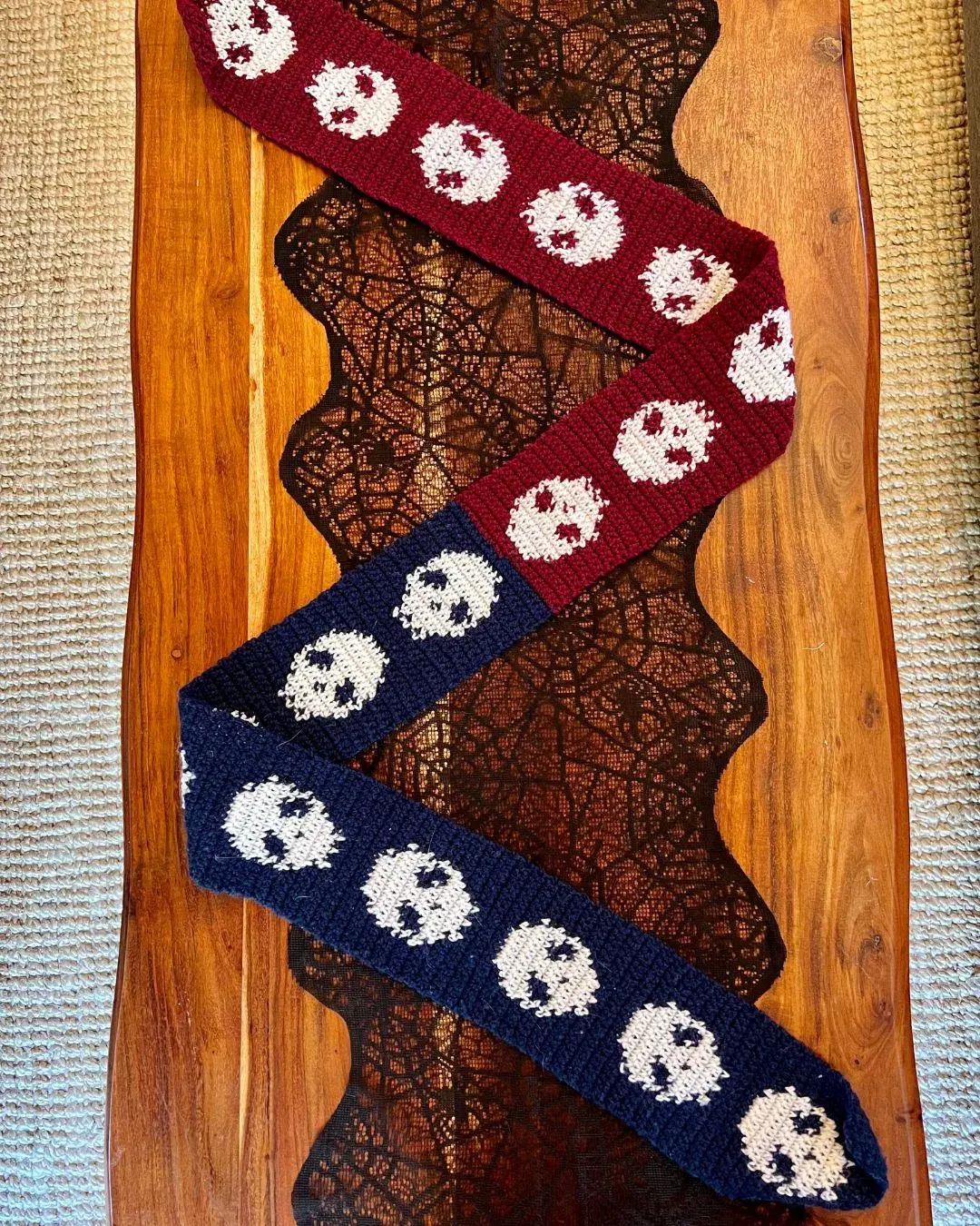 a scarf half blue and half red with repeating skulls for the whole length of the scarf laid on a table. The skulls change direction at the midpoint of the scarf so that they are facing up while wearing