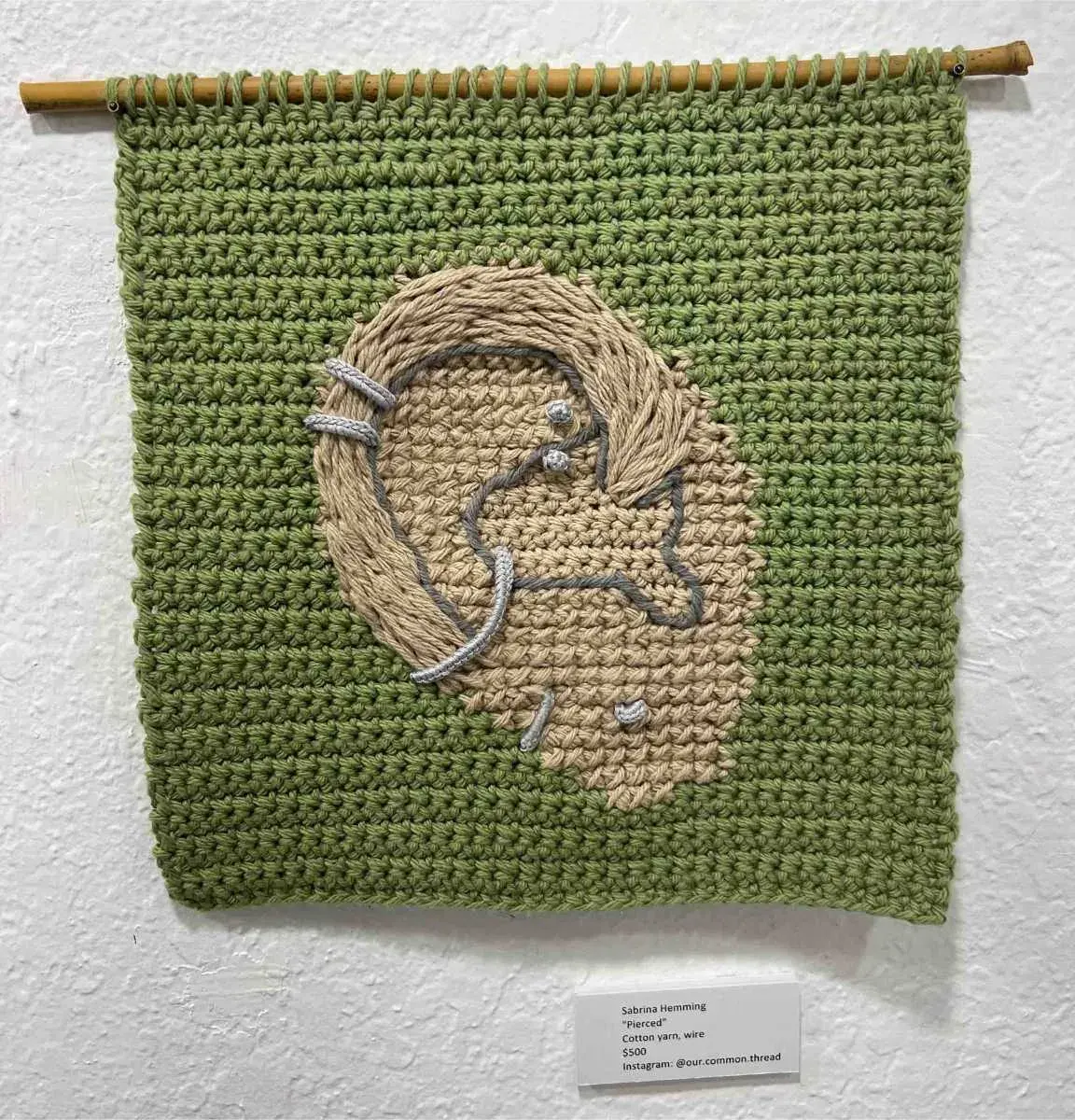 a crochet tapestry with a light breen background. There is an ear defined using embroidery and crochet, with crocheted piercings going through the tapestry.