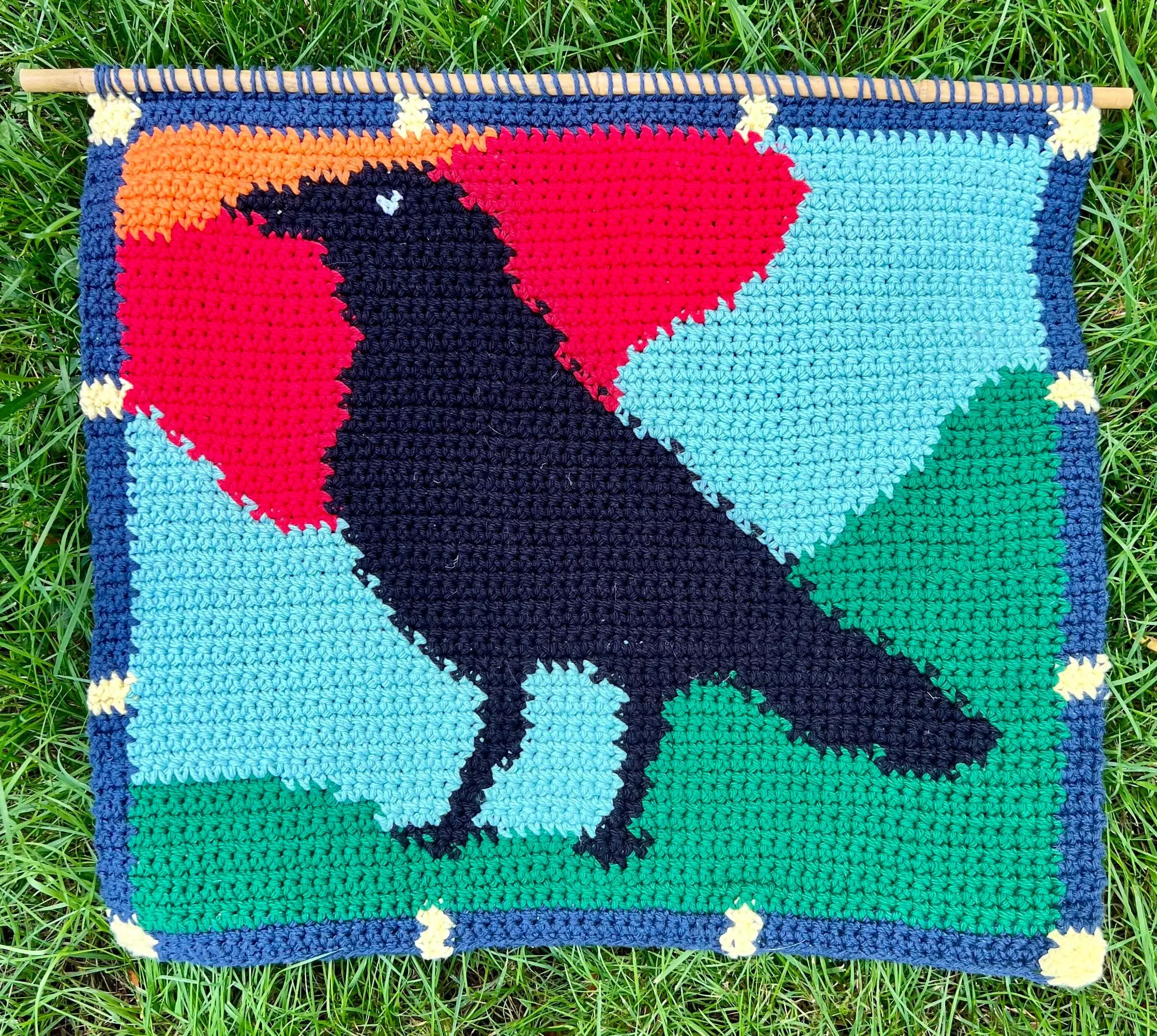 a crochet tapestry with a side-profile crow taking up most of the frame. The background is an abstracted green field and blue sky with a sunset approaching. Colors are framed around the crow similarly to how pieces of stained glass are placed