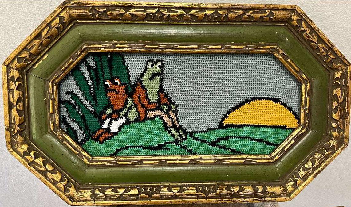 a crochet tapestry with frog and toad in the left forefront back to back, some foliage behind them. To the right is a sunset over the hills. Tapestry is in an ornate green frame with gold detailing