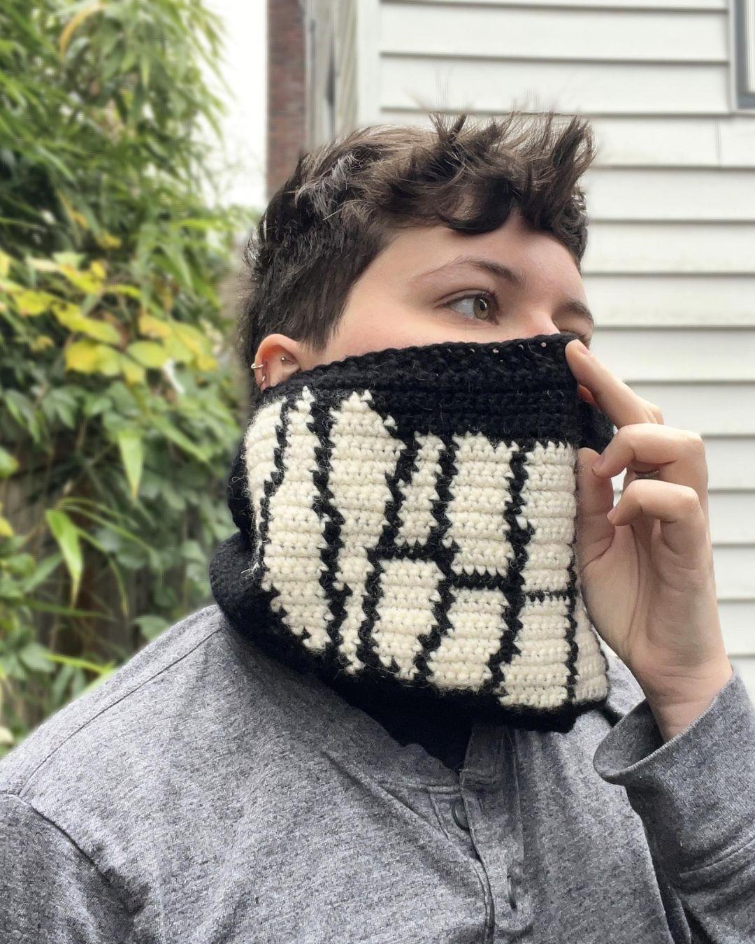 person with short brown hair wearing a crocheted cowl held up  over nose standing outside with a blurred background of greenery and  a white building. The cowl is black with white smiling teeth across  the front and 3 sets of fangs on the sides