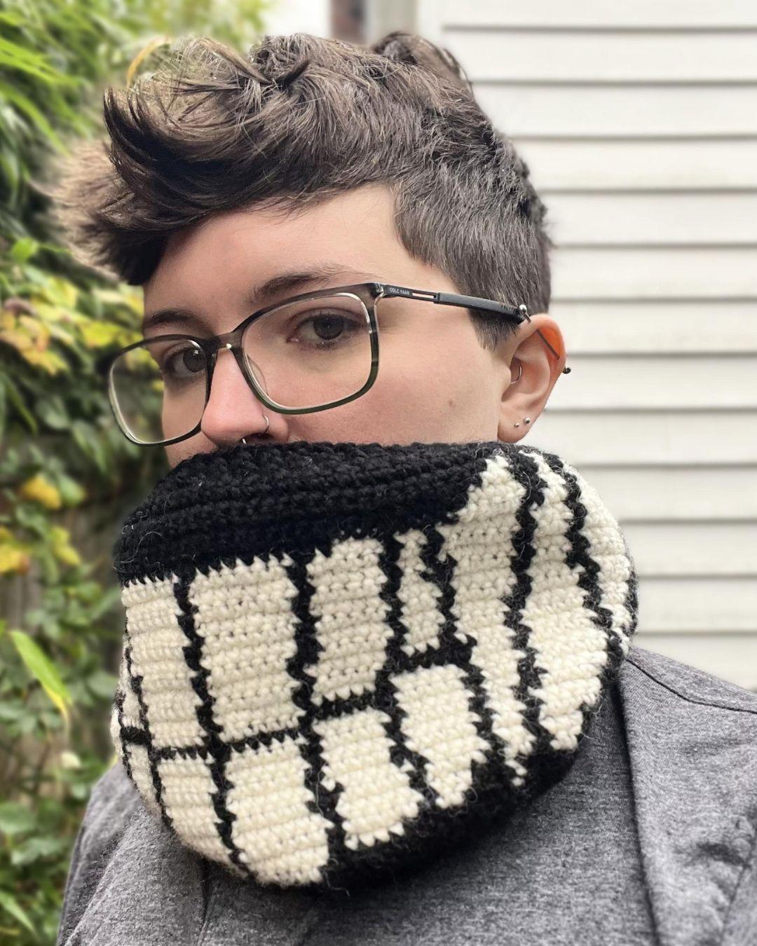 person with short brown hair wearing a crocheted cowl standing outside with a blurred background of greenery and a white building. The cowl is black with white smiling teeth across the front and 3 sets of fangs on the sides