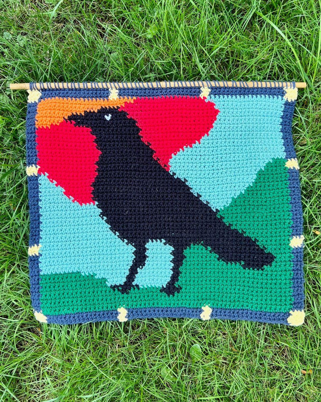 a crochet tapestry with a side-profile crow taking up most of the frame. The background is an abstracted green field and blue sky with a sunset approaching. Colors are framed around the crow similarly to how pieces of stained glass are placed