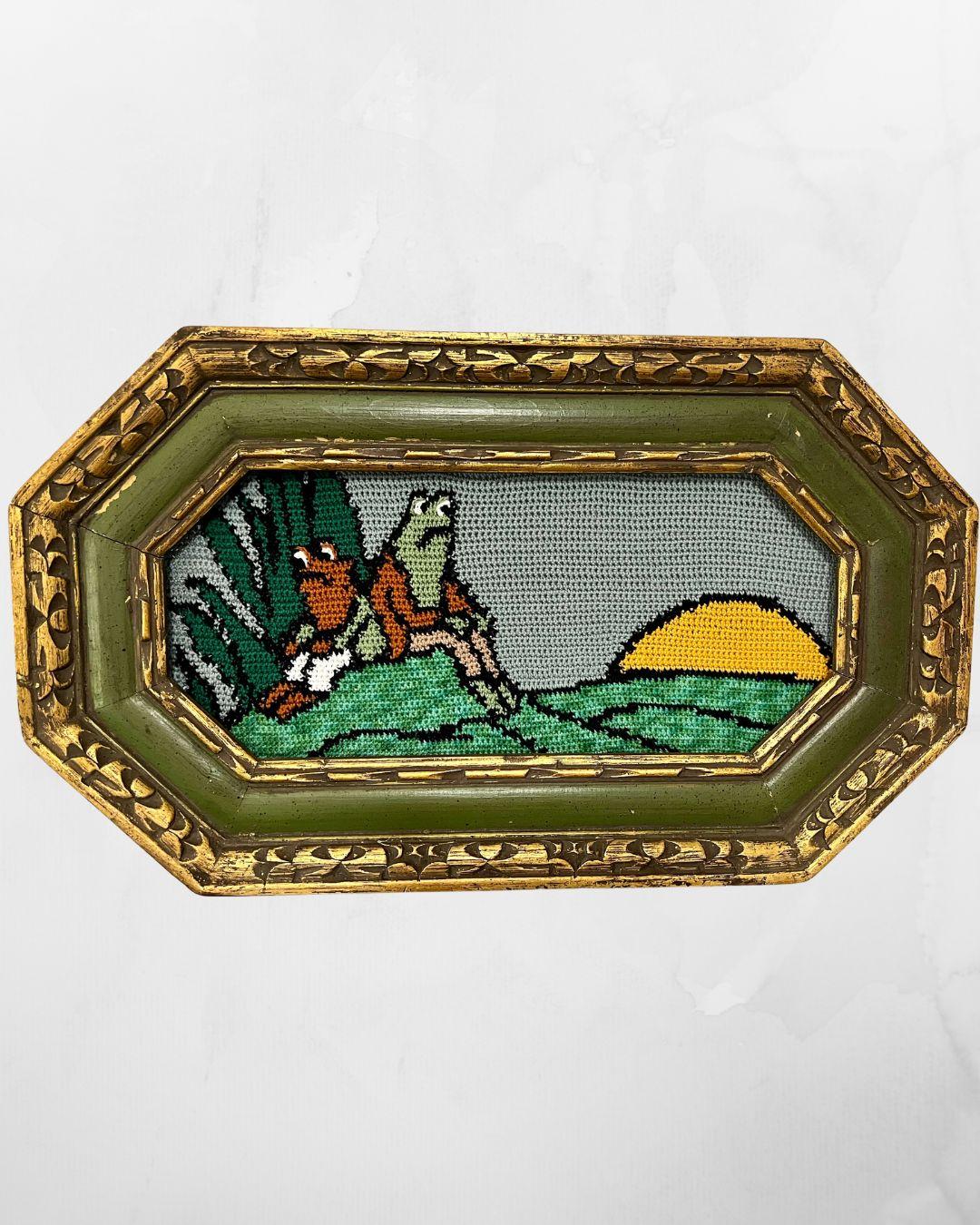 a crochet tapestry with frog and toad in the left forefront back to back, some foliage behind them. To the right is a sunset over the hills. Tapestry is in an ornate green frame with gold detailing