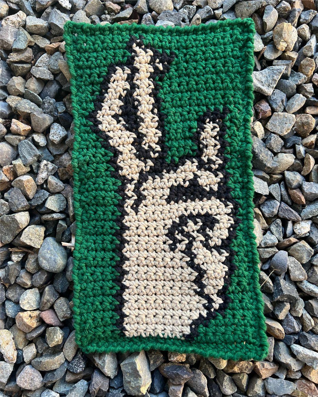 a crochet tapestry placed on gravel with a green background and a side view of a hand with crossed fingers