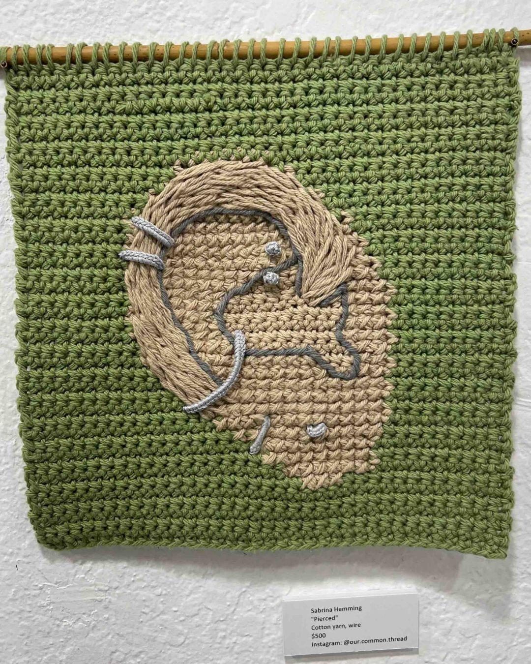 a crochet tapestry with a light breen background. There is an ear defined using embroidery and crochet, with crocheted piercings going through the tapestry.